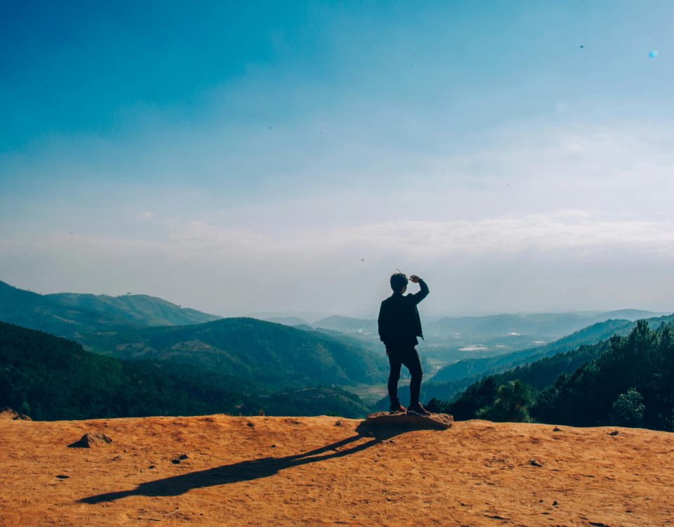A lone traveler stands on a hilltop, gazing over a vast mountain landscape under a clear blue sky.