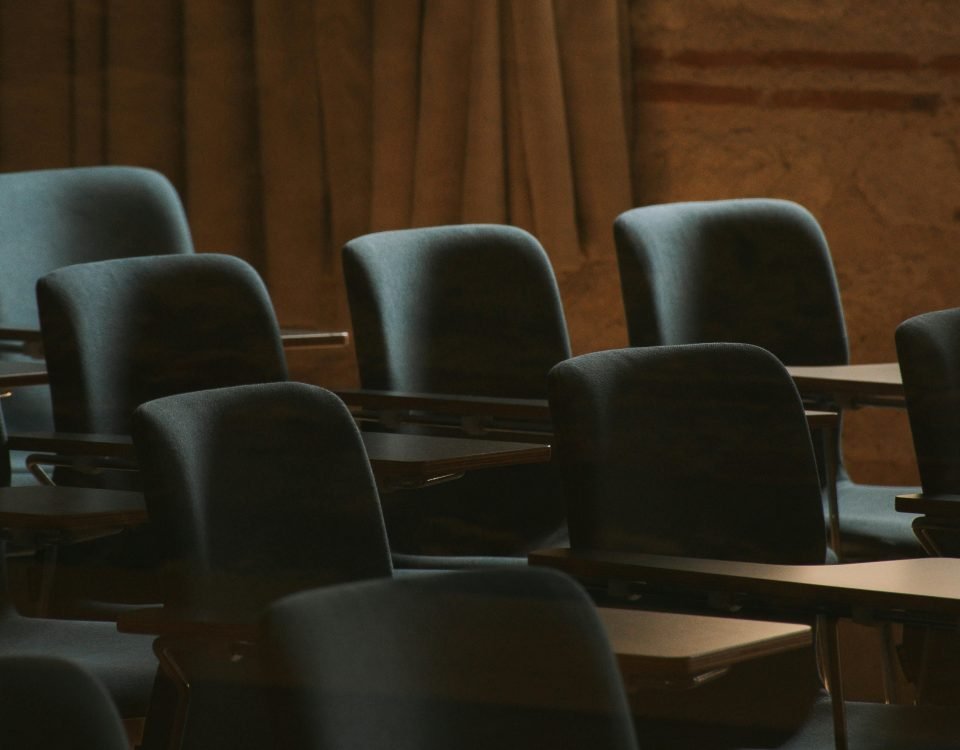 A dimly lit empty classroom with multiple rows of chairs and tables, suggesting a moment of calm before or after class.