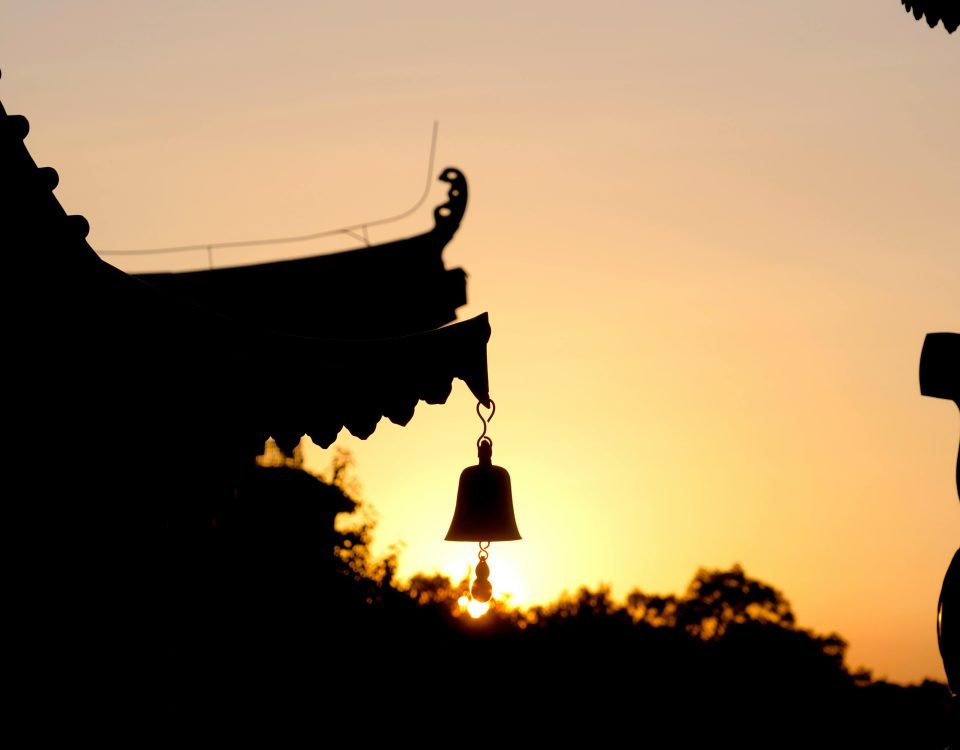 Silhouette of a bell hanging from a rooftop against a vibrant sunset background.