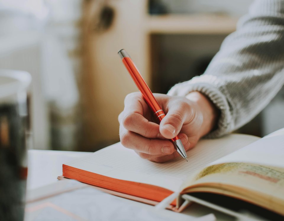 A person writing in a notebook with a red ballpoint pen, showcasing detail and focus.